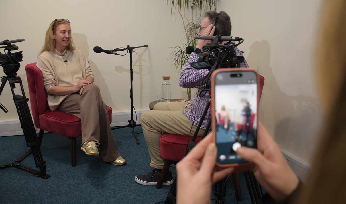 A photograph showing a women being interviewed by a man. They are both sat down. There is camera equipment in shot to the left and right of frame. The photo is take over the shoulder of a women taking a photo on her phone.